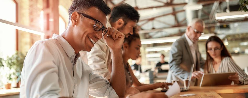 A man smiles while reading a laptop in an open office space with coworkers.